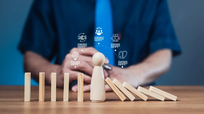 Wooden figure stands firm as dominoes fall, symbolizing resilience and stability in business crisis. Icons above depict innovation, teamwork, and decision-making under pressure