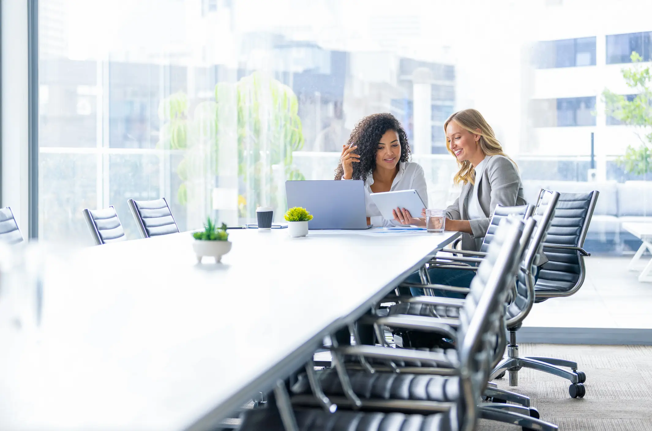 Business colleagues having a conversation in a board room. They are both young business people casually dressed in a modern office. Could be an interview or consultant working with a client. She is listening and smiling. They are using a laptop and digital tablet. Mixed ethnic group. There is a large window behind them